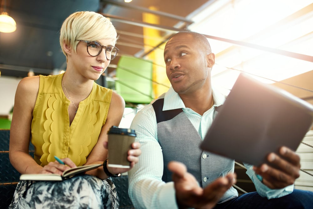 Two creative millenial small business owners working on social media strategy using a digital tablet while sitting in staircase Two creative millenial small business owners working on social media strategy using a digital tablet while sitting in staircase